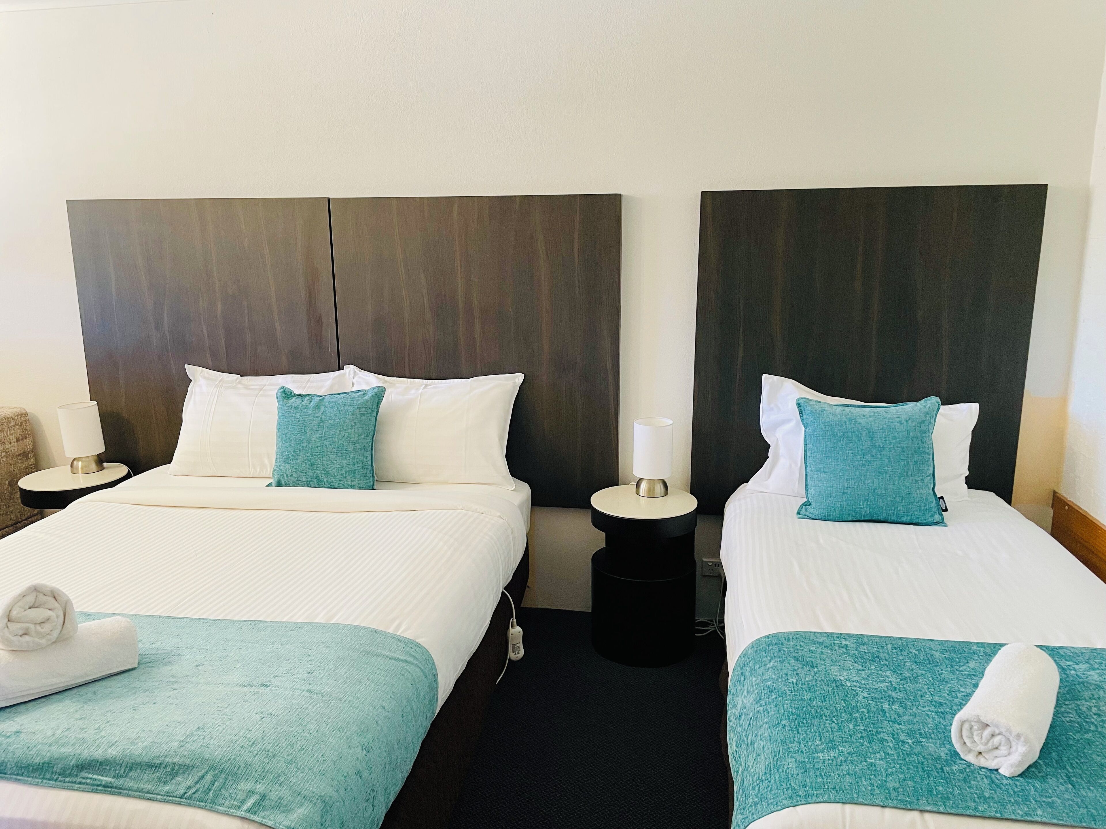 Two double beds with white linens and teal accents, a dark wood headboard, a small bedside table with a lamp, and a rolled towel on each bed.