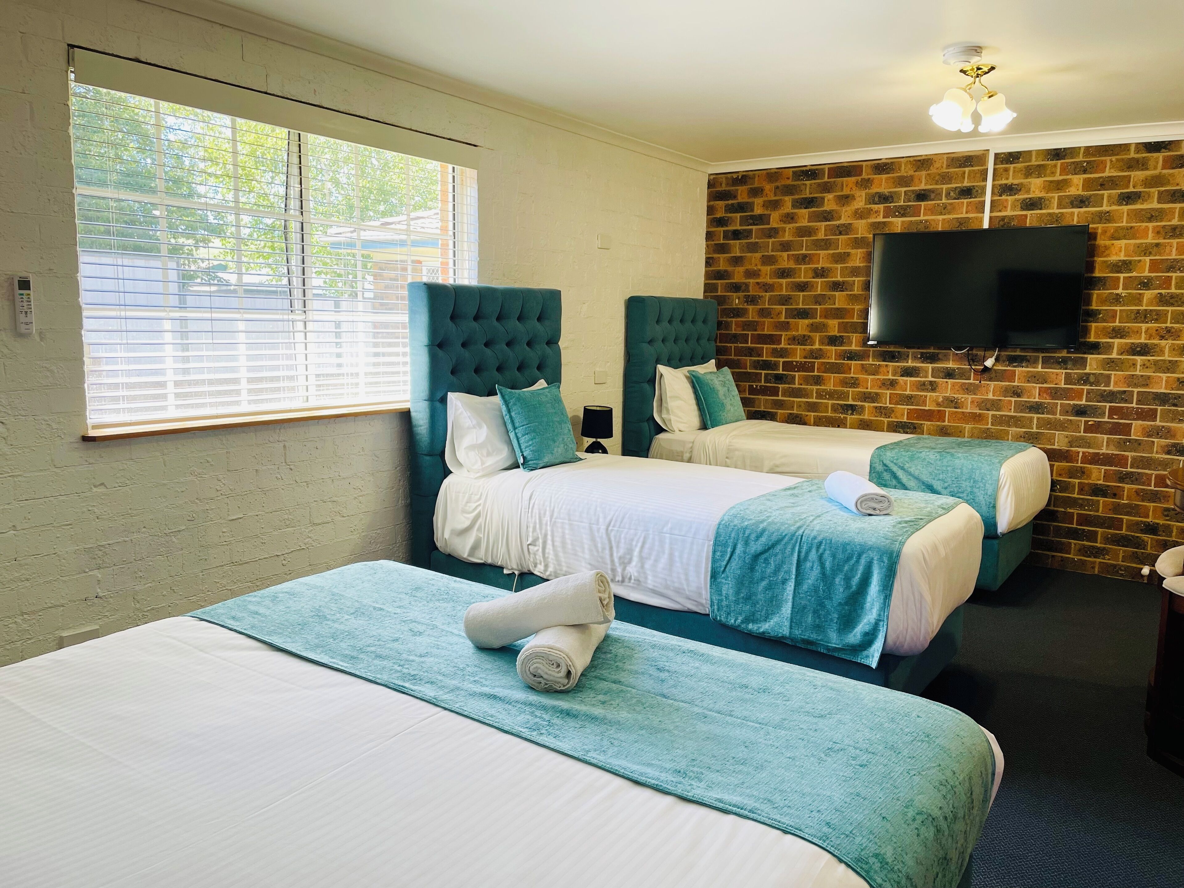 Une chambre d’hôtel avec deux lits, un téléviseur fixé au mur en briques et une fenêtre avec des stores.