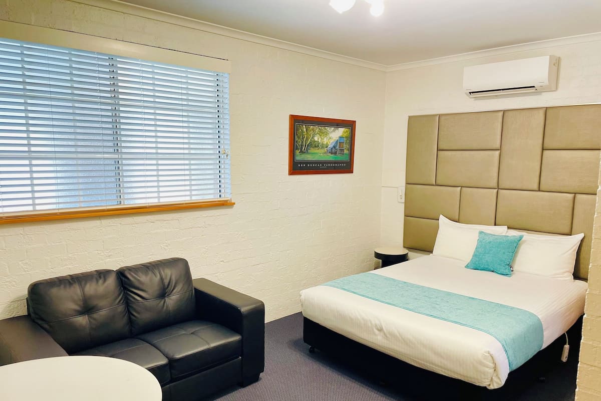 Habitación de hotel con cama, sofá, mesa redonda, ventana con persianas, unidad de aire acondicionado y un cuadro en la pared.