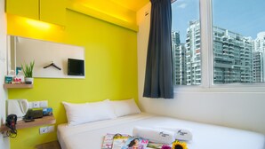 A hotel room with a bed, a mirror, a window with a curtain, a shelf with toiletries, and a magazine on the bed.