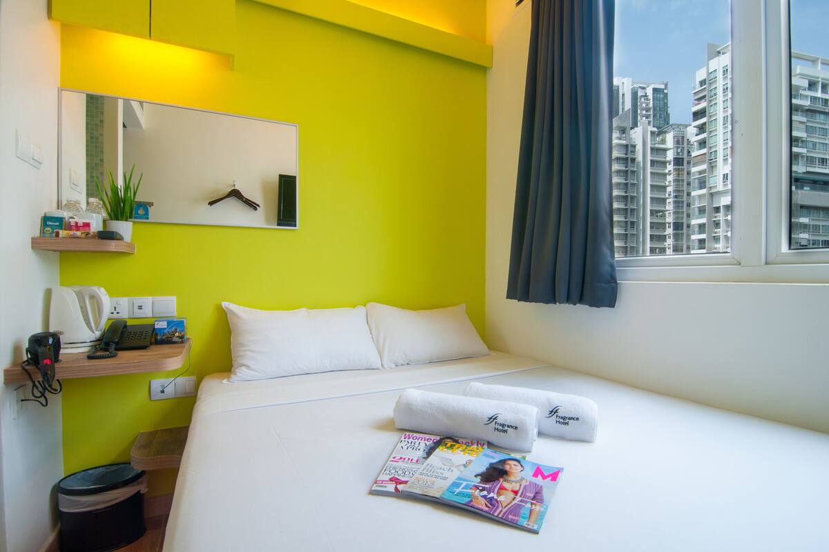 A hotel room with a bed, a mirror, a window with curtains, a shelf with toiletries, and a magazine on the bed.