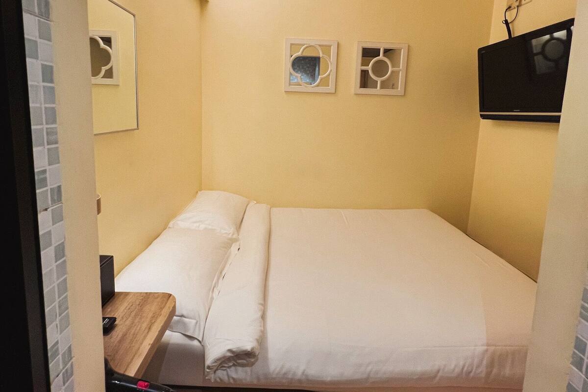 Une petite chambre avec un lit simple, un bureau en bois, une chaise et un téléviseur fixé au mur.