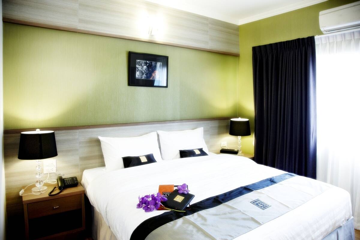 A hotel room with a bed, two bedside tables, a nightstand, a telephone, a lamp, a picture on the wall, and a curtain.