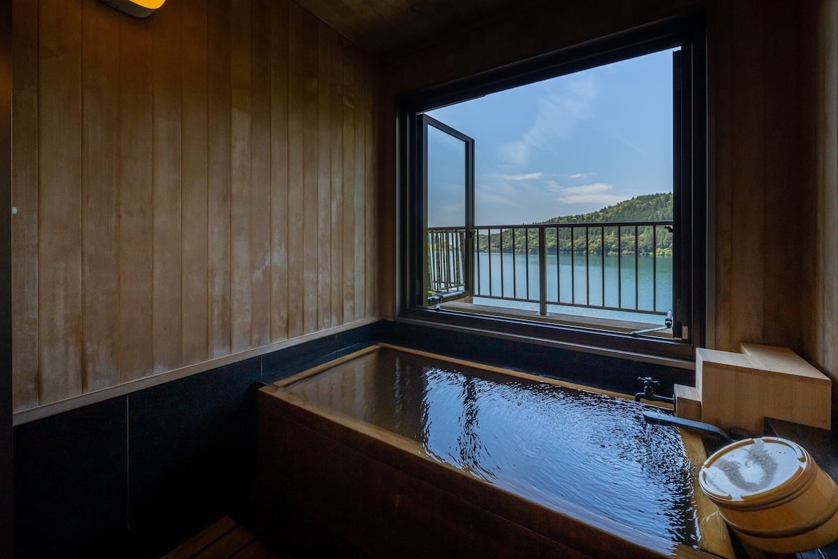 A wooden interior with a large bathtub, a window with a view of a lake and hills, and a wooden bench.