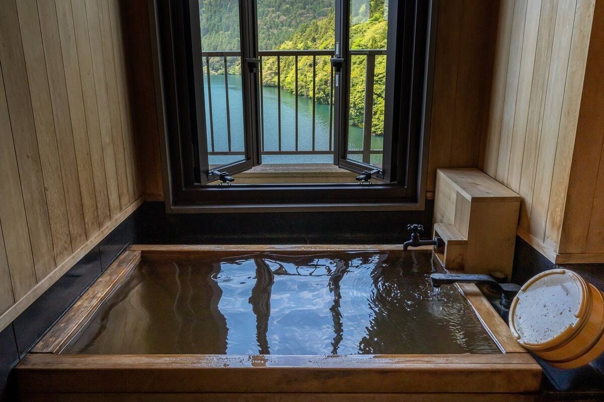 A wooden bathtub with a view of a lake and forest through a large window.
