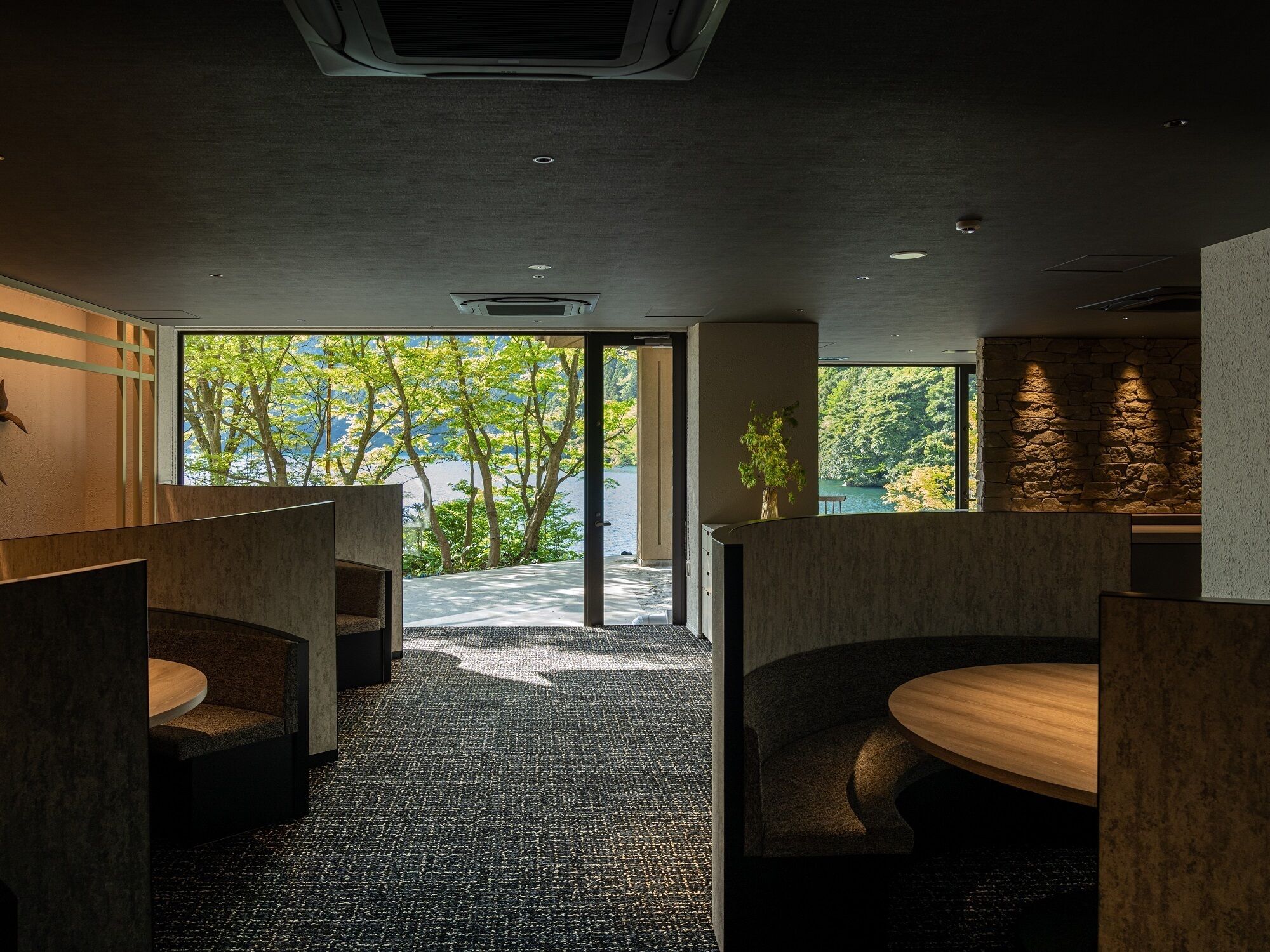 A modern hotel lobby with a view of a lake and trees. There are seating areas with wooden tables and chairs.