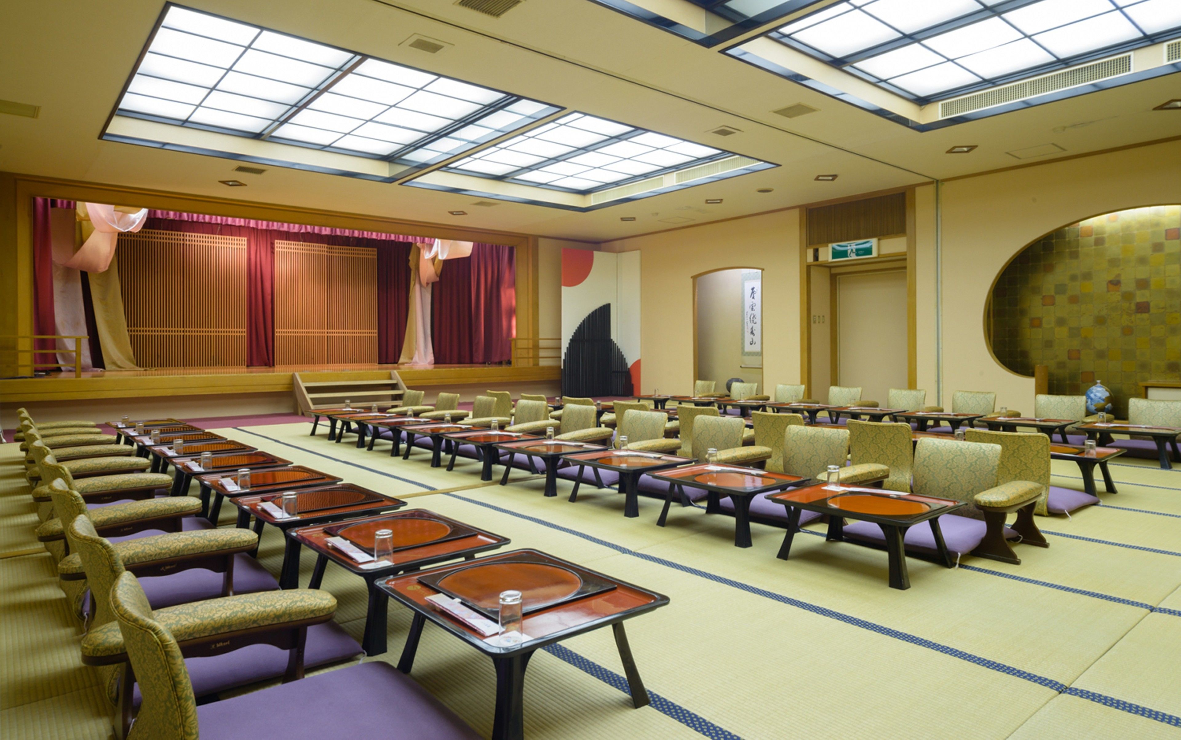 Un salón de conferencias con filas de mesas y sillas, un escenario con cortinas y un gran ventanal con tragaluces.