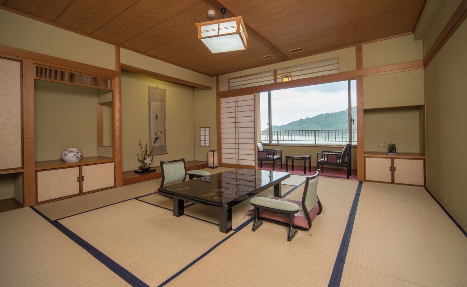 A traditional Japanese room with tatami flooring, a low table, chairs, and a view of a mountain.