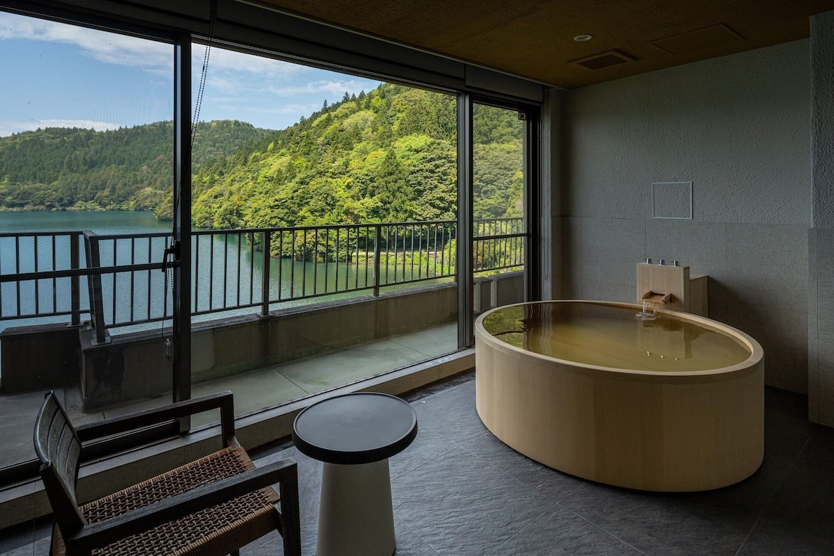 A modern bathroom with a large bathtub, a view of a lake and forest, and a seating area with a chair and table.