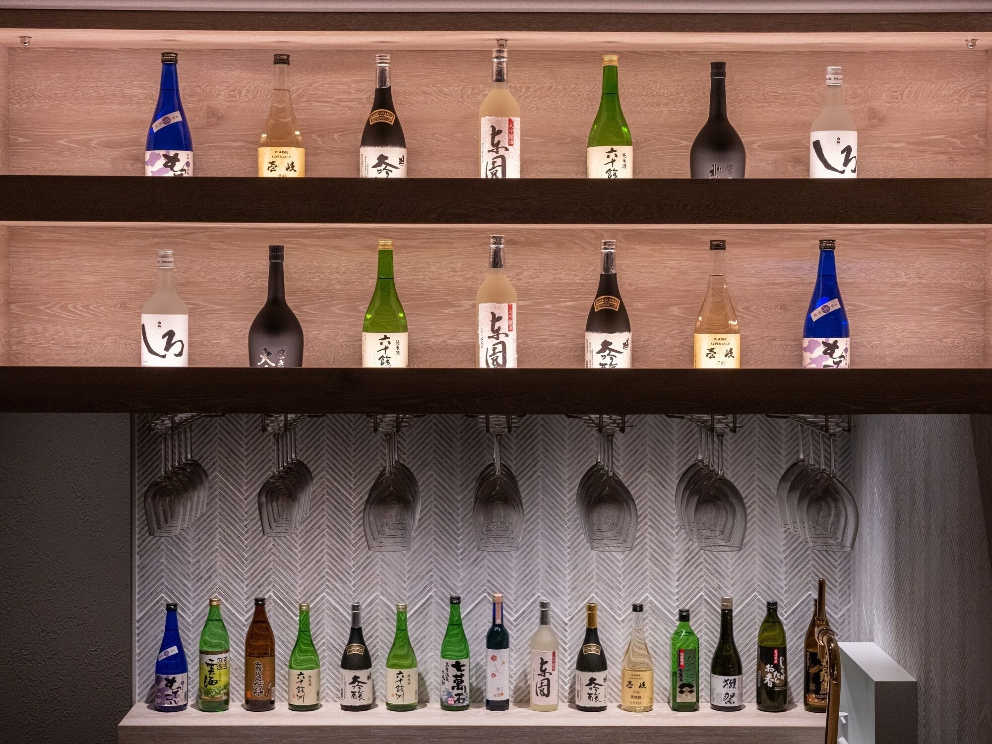 A selection of sake bottles on wooden shelves with a herringbone patterned wall in the background.