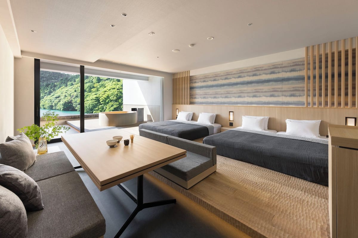 A modern hotel room with a large window offering a view of a forested area. There is a sofa, a small table with bowls, and two beds with pillows.