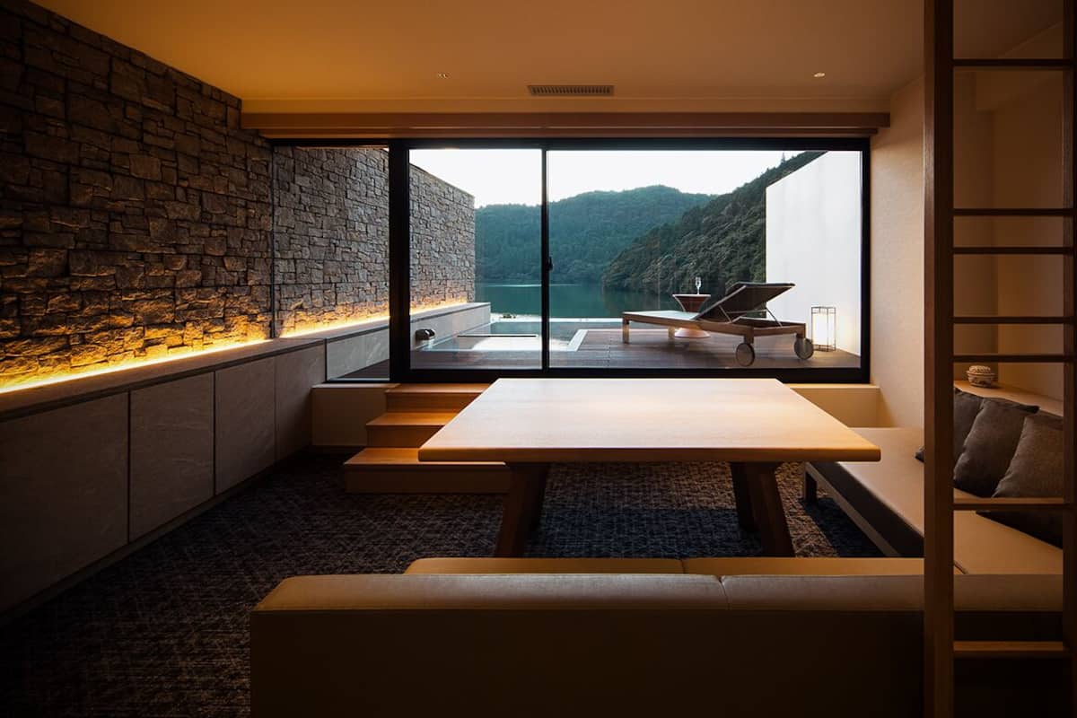 A modern interior with a stone wall, a large window overlooking a lake, and a seating area with a table and chairs.