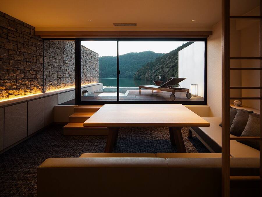 Um interior moderno com uma parede de pedra, uma janela grande com vista para um lago e uma área de estar com mesa e cadeiras.