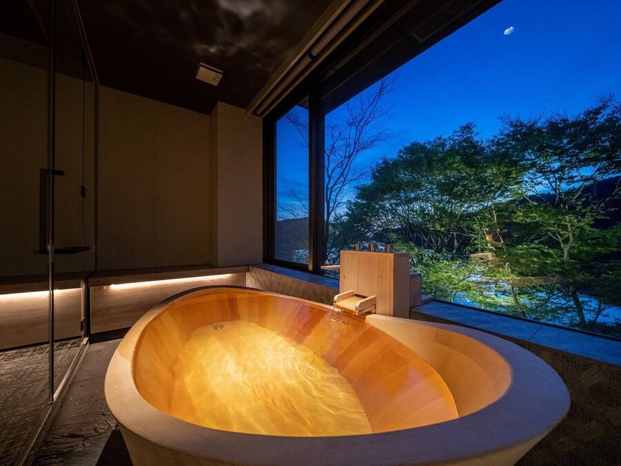 A modern bathroom with a wooden bathtub, large windows, and a view of trees and a body of water.