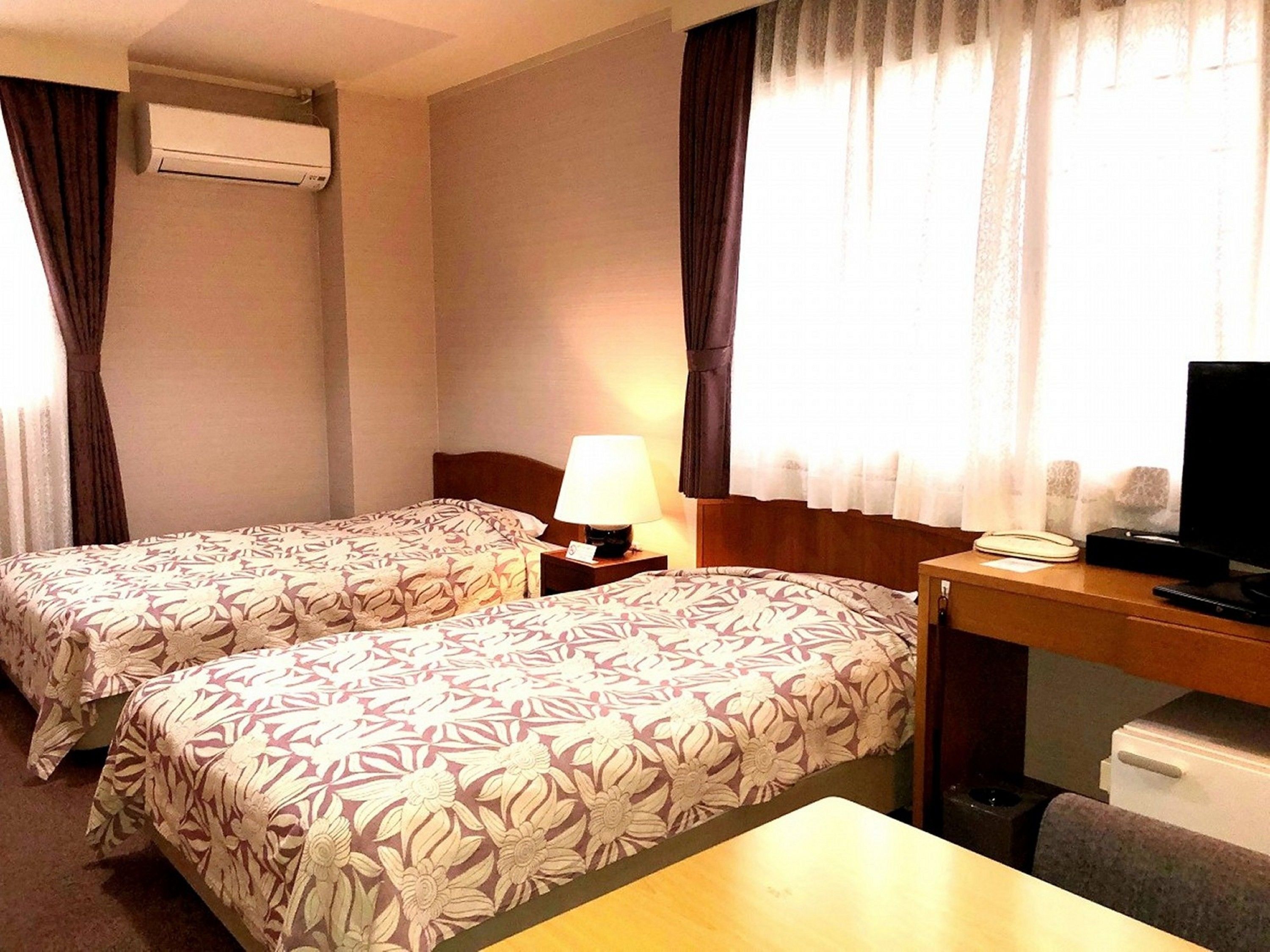 A hotel room with two beds, a desk, a television, and an air conditioning unit.