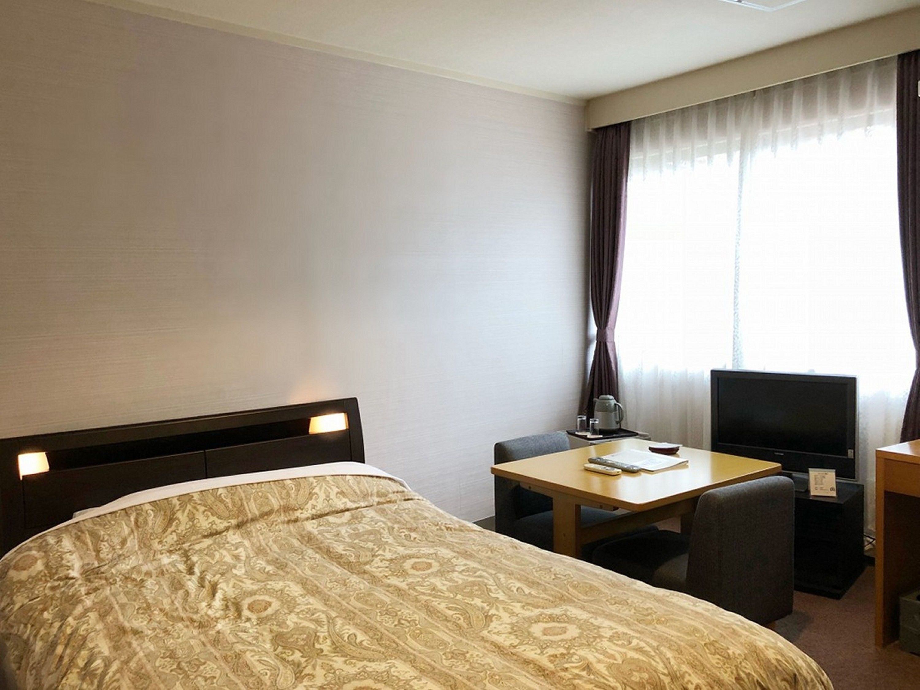Quarto de hotel com uma cama grande, uma escrivaninha com televisão e uma janela com cortinas.