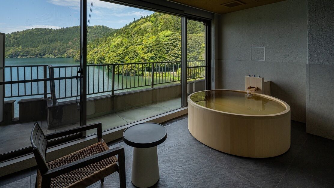 A room with a large window, a wooden bathtub, a bench, and a small table.