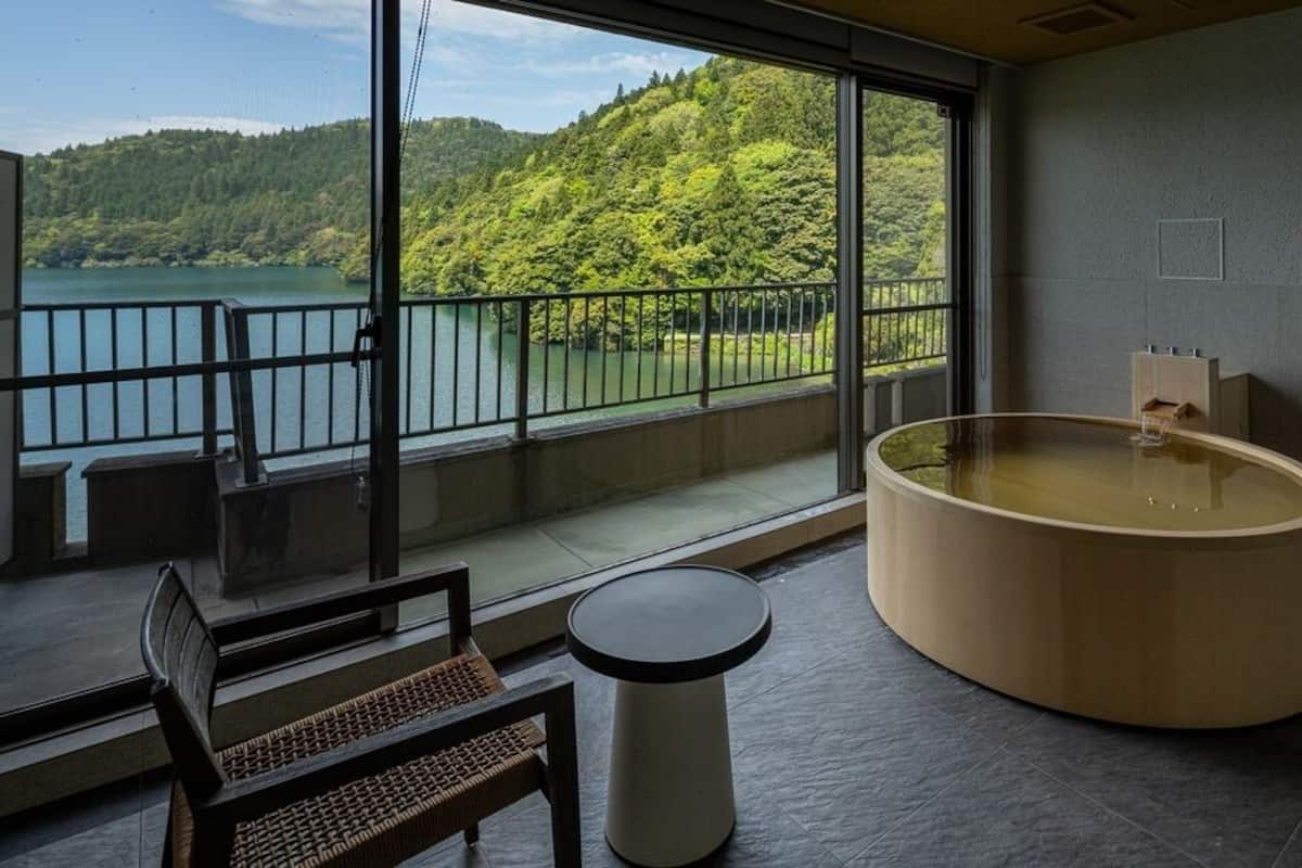 A room with a large window, a wooden bathtub, a bench, and a small table.