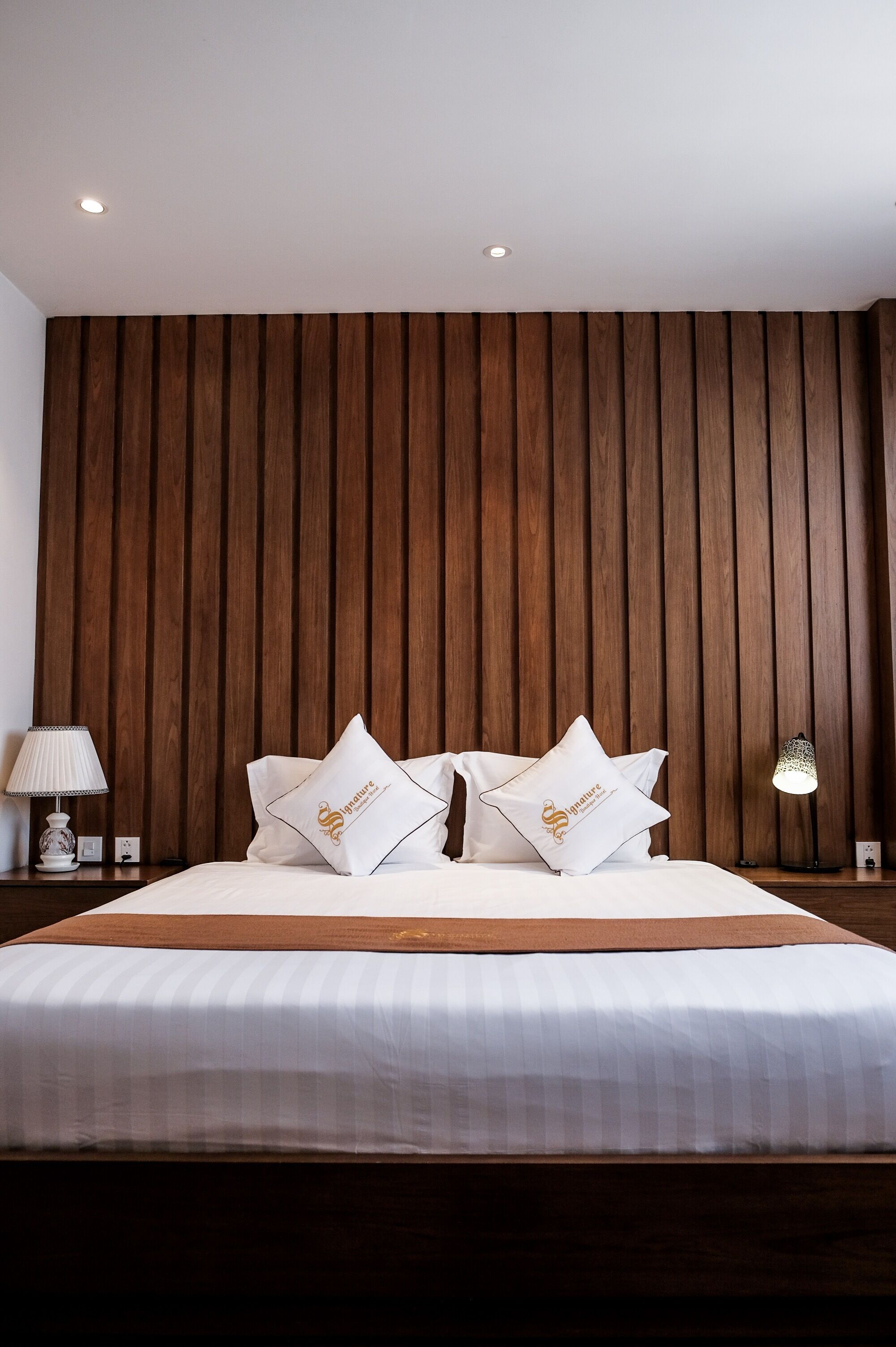 Una cama bien hecha con ropa blanca y dos almohadas, cabecera de madera, lámpara de noche y aplique de pared.