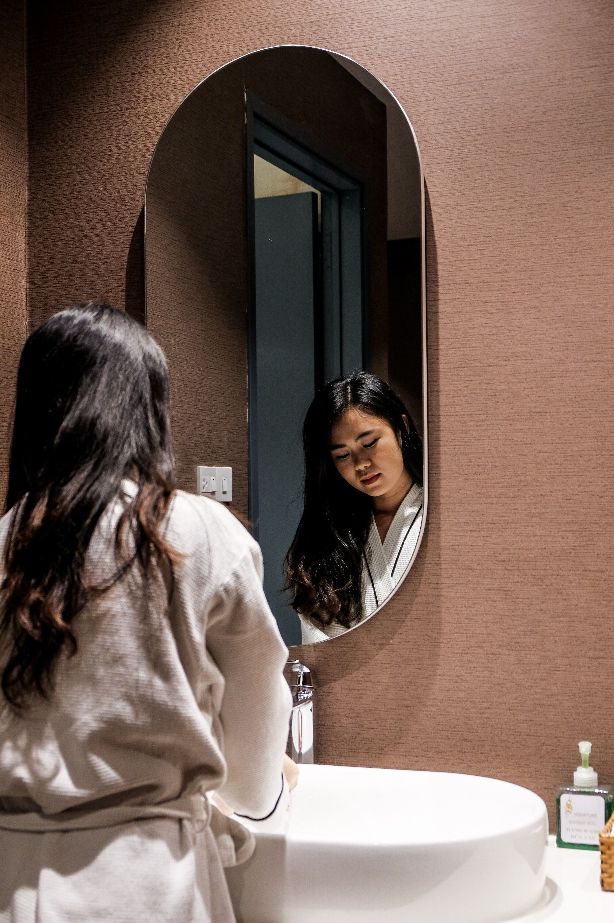 Une personne vêtue d’une robe se tient devant un miroir de salle de bain, devant un lavabo blanc, et une bouteille de savon liquide est posée sur le comptoir.