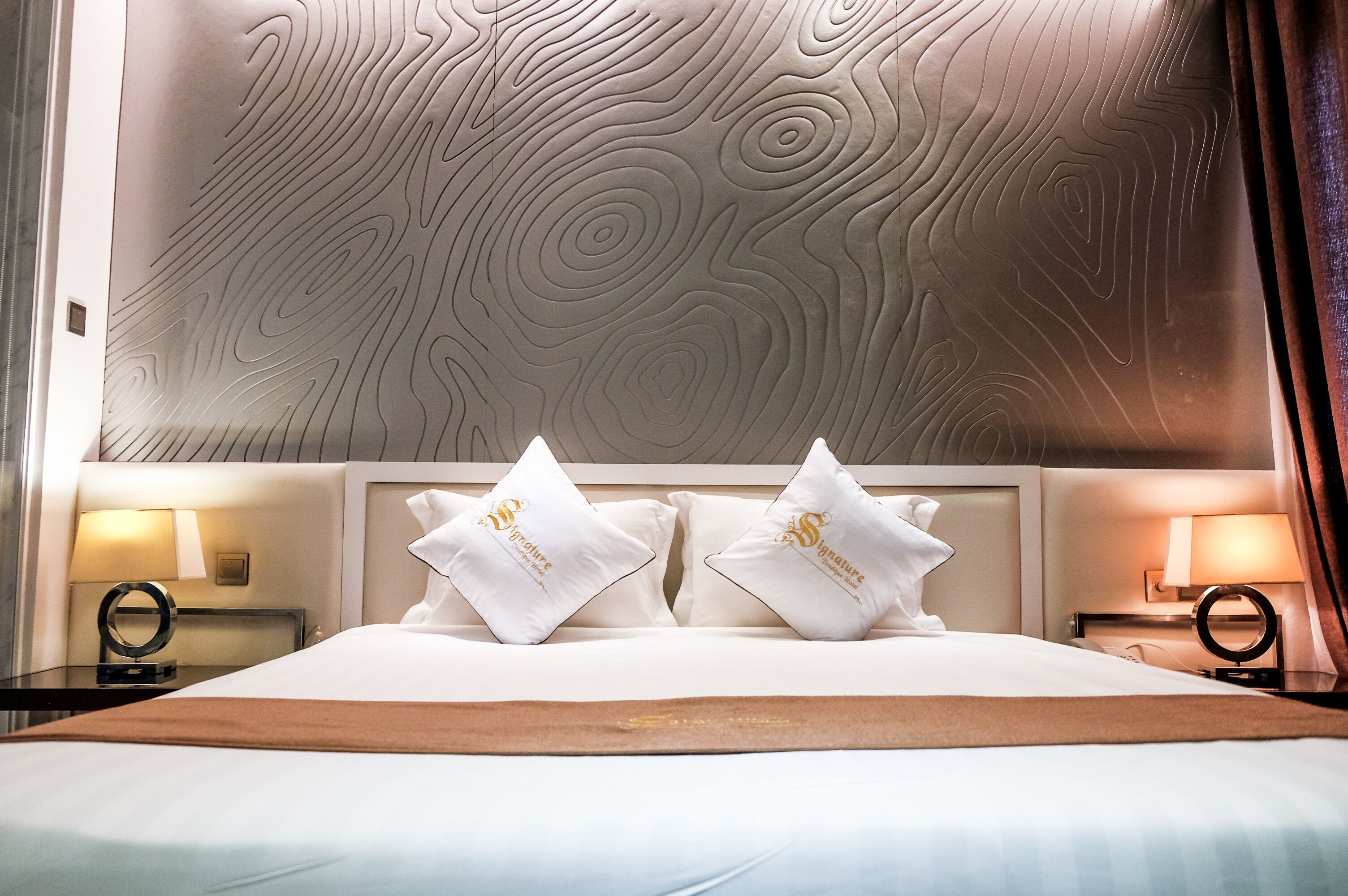A hotel room with a large bed, two white pillows with a logo, a headboard with a textured design, and bedside lamps.