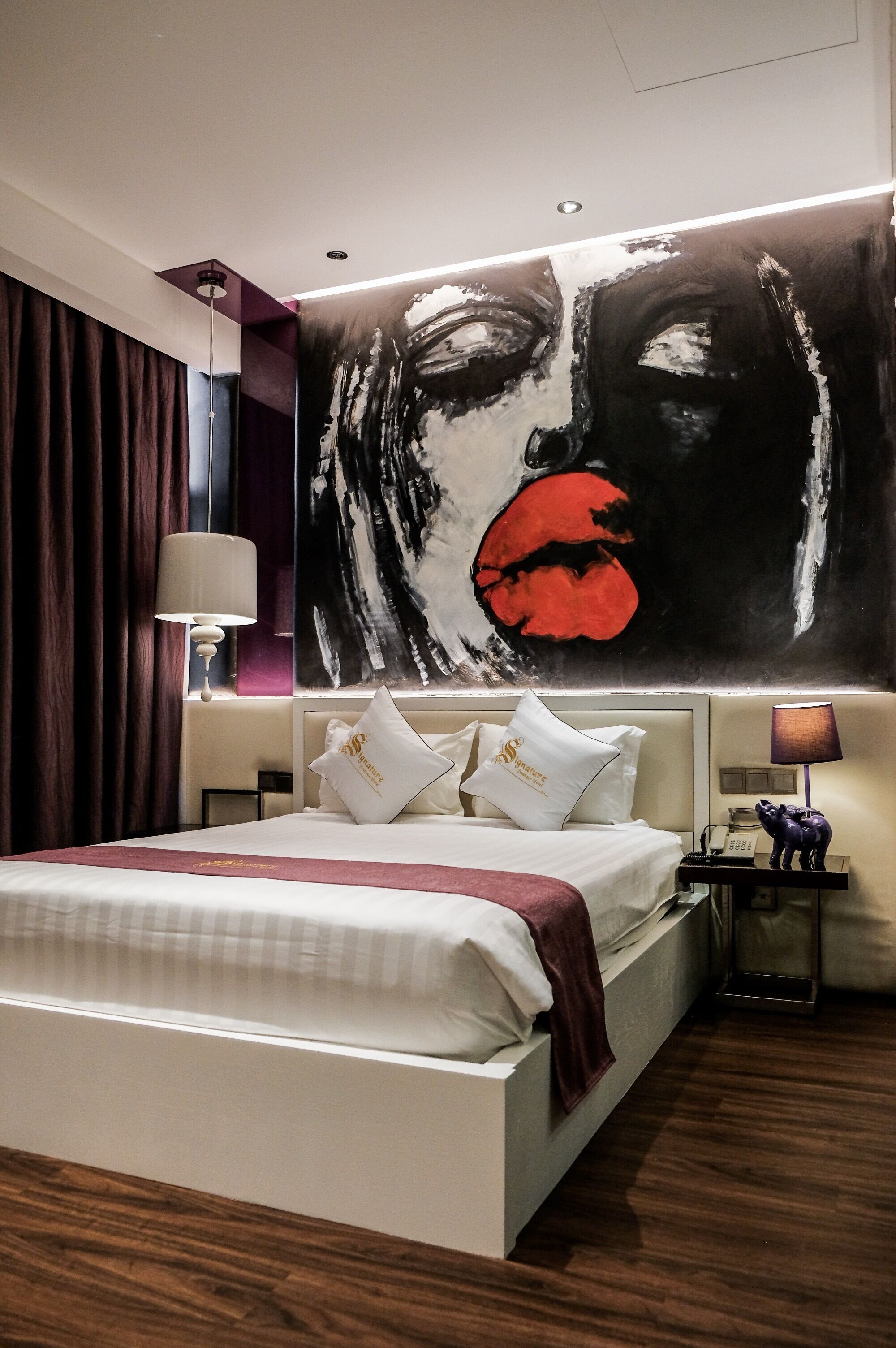 A modern hotel room with a large bed, a nightstand with a lamp, and a wall painting.