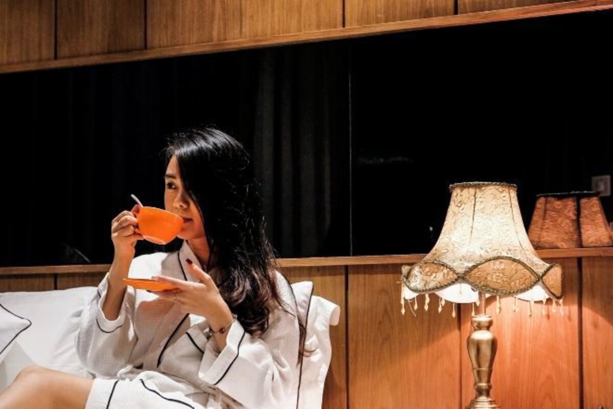 Een persoon in een witte badjas zittend op een bed met een houten hoofdeinde, een oranje beker vasthoudend. Er staat een lamp met een gematteerde lampenkap op een nachtkastje naast het bed.
