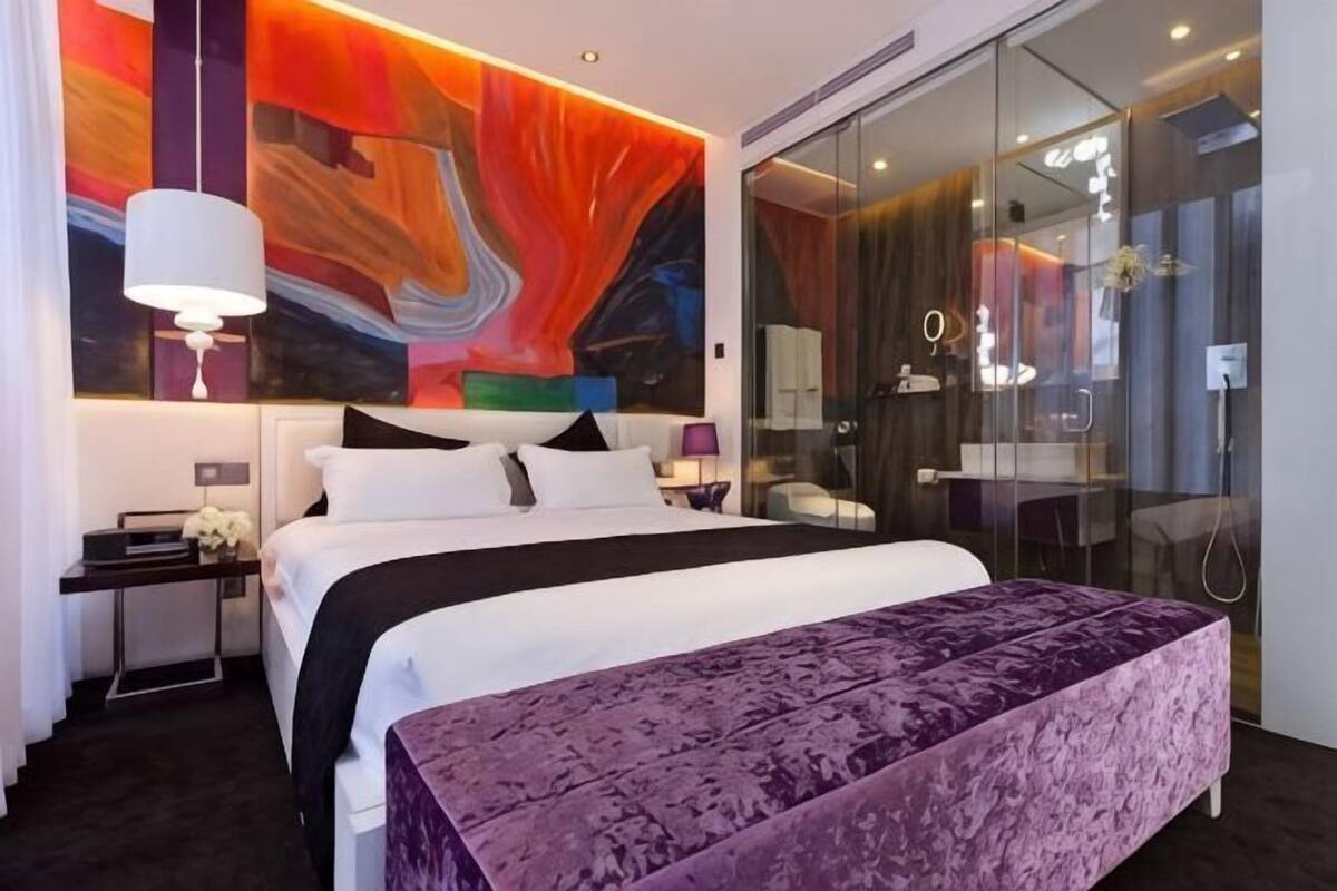 Una habitación de hotel moderna con una cama grande, una mesita de noche y un box de ducha de vidrio, además de un vibrante mural abstracto en la pared.