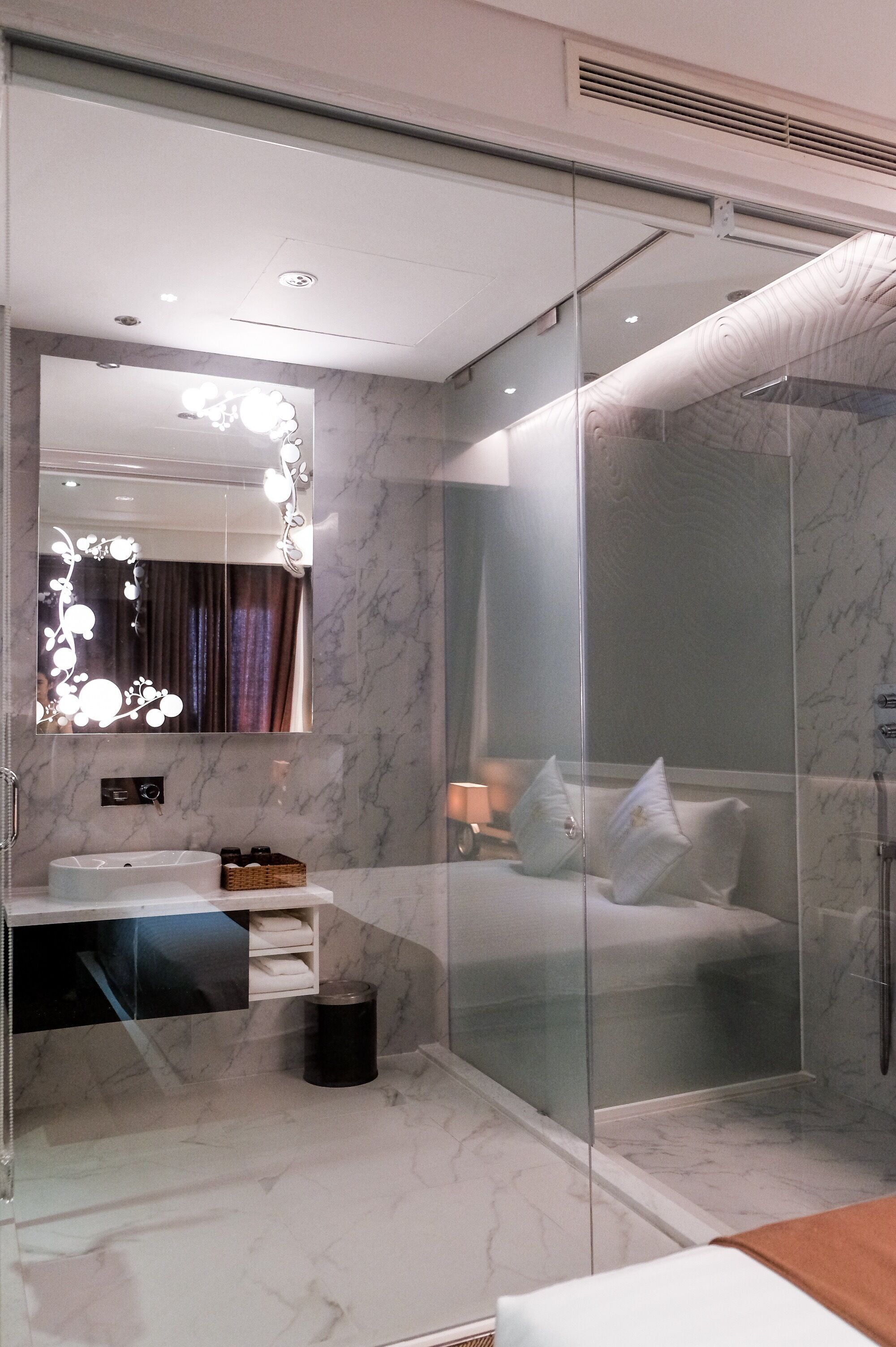 Un baño moderno con ducha envuelta en vidrio, tocador con espejo y cama con ropa de cama blanca.