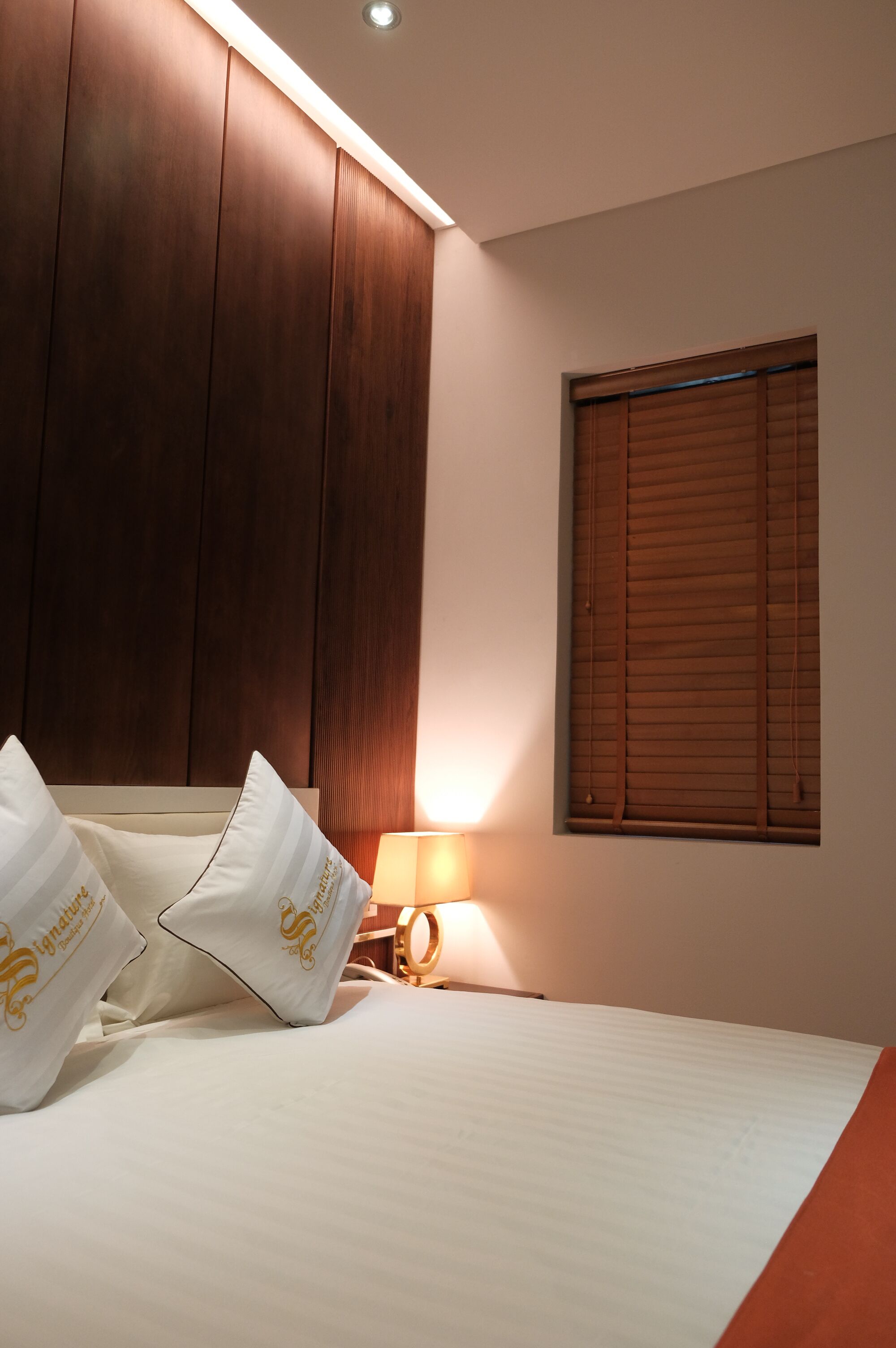 Una habitación de hotel con cama, almohadas, mesita de noche, lámpara y ventana con persianas.