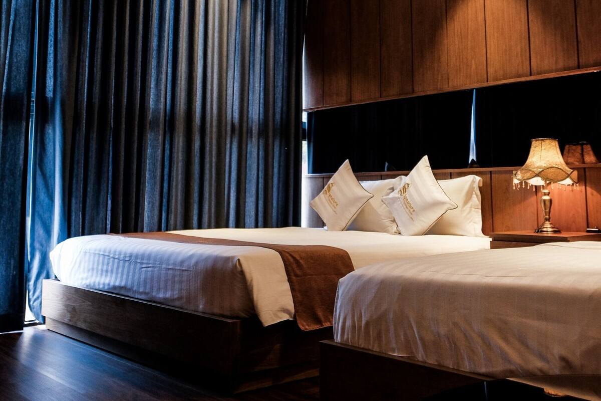 Habitación de hotel con dos camas, paneles de madera, un ventanal con cortinas y una mesita de noche con lámpara.