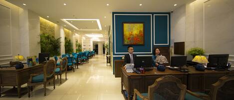 A hotel lobby with a reception desk, several chairs, a large painting, and a chandelier.