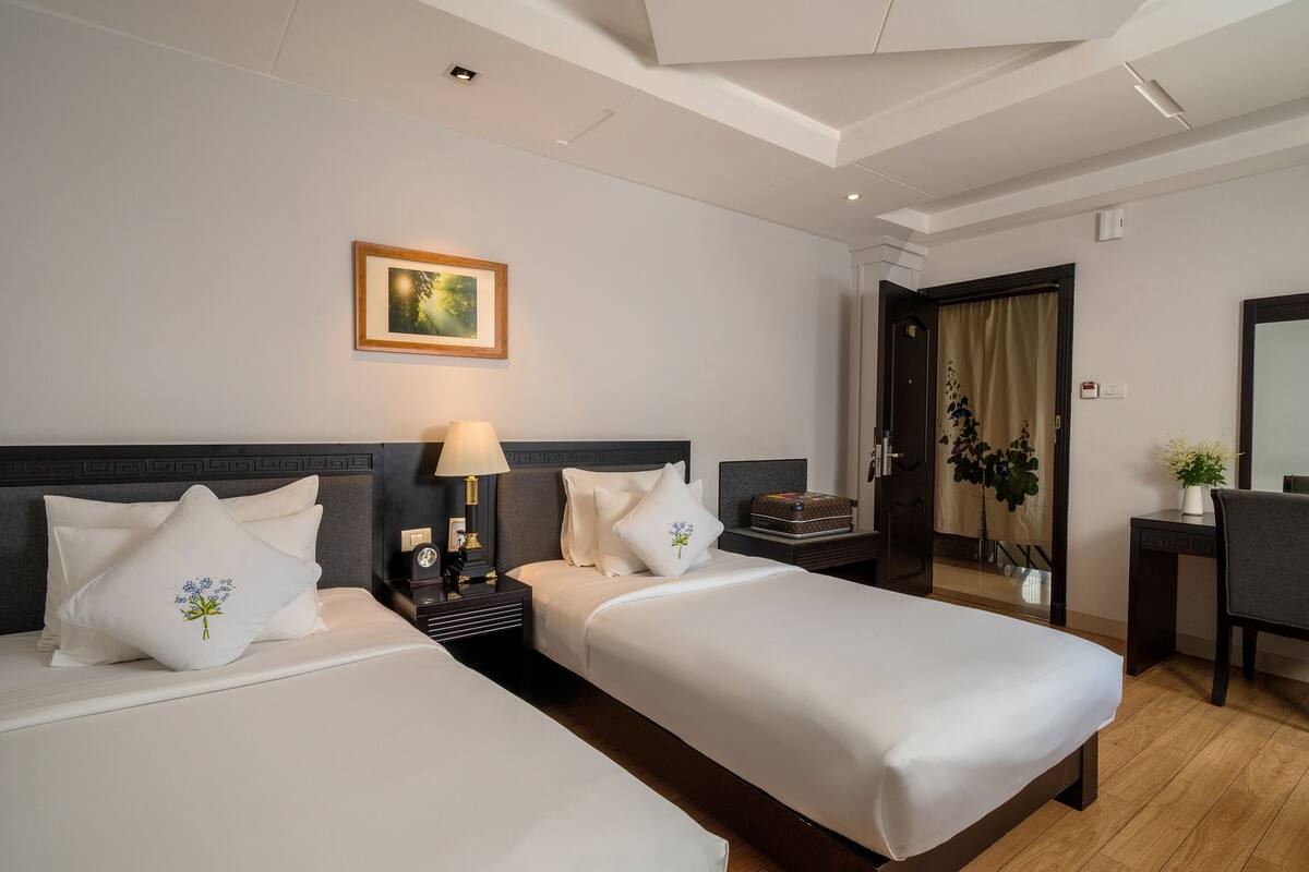 Een hotelkamer met twee bedden, een nachtkastje met een lamp, een lijst met een afbeelding aan de muur, en een bureau met een stoel.