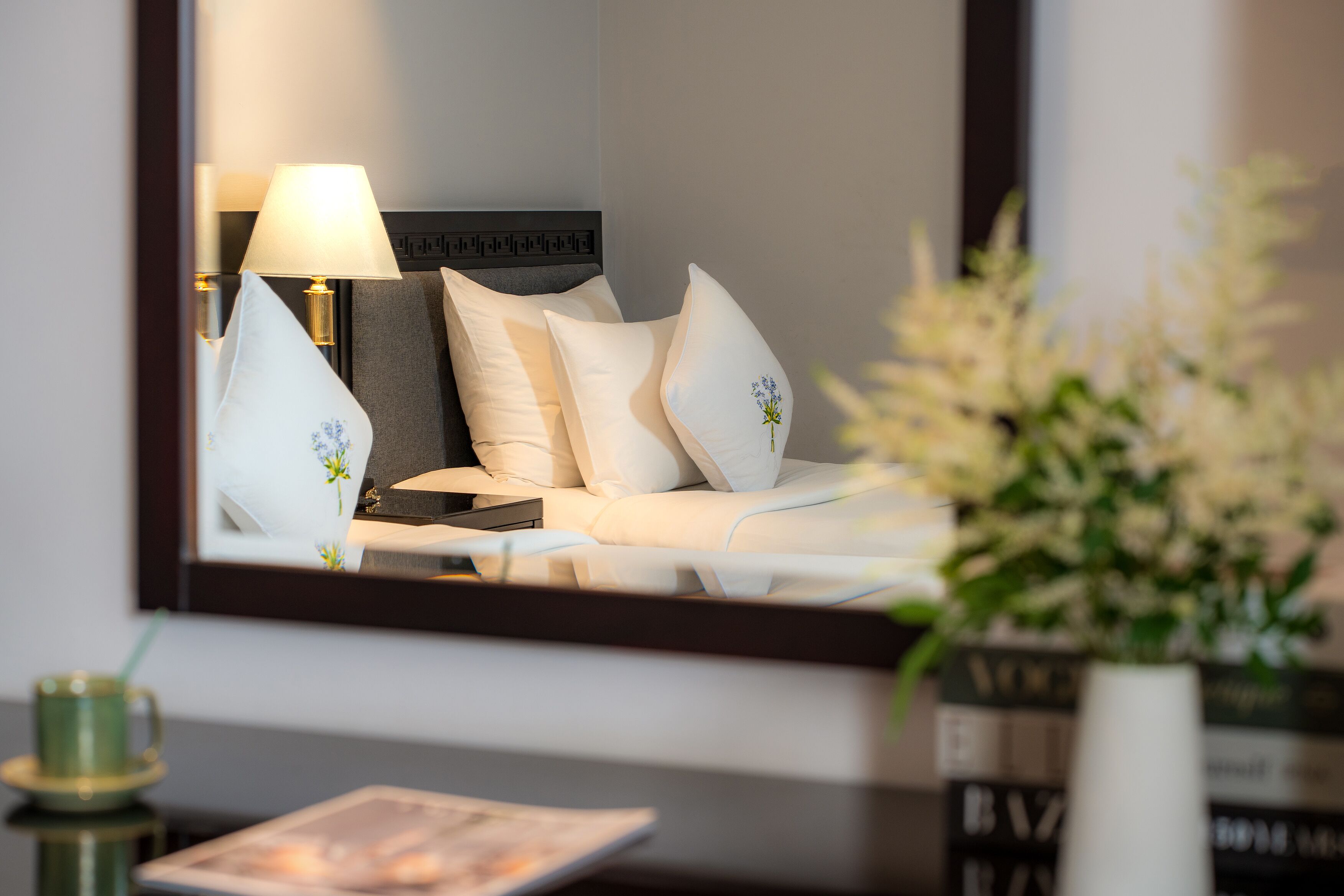 Une chambre d’hôtel avec un lit, des oreillers, une table de chevet avec une lampe, un vase de fleurs et un magazine sur la table.