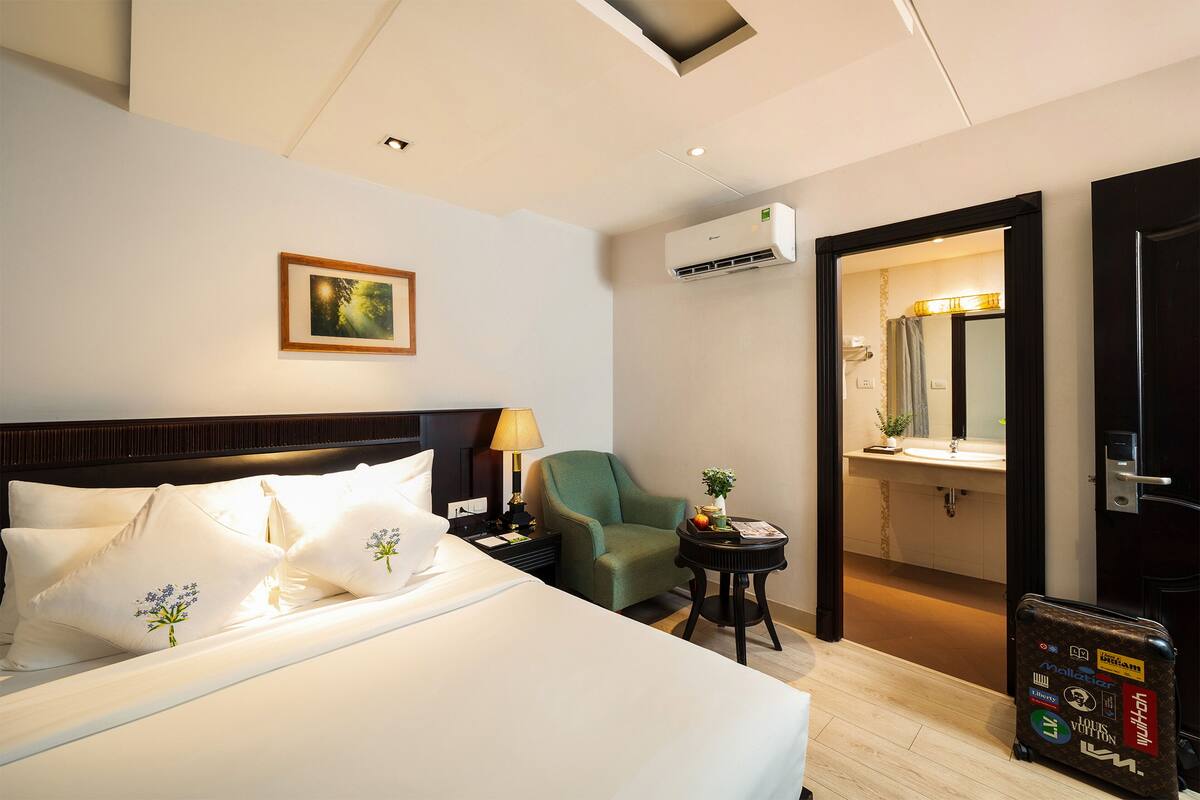 Een hotelkamer met een bed, kussens, een nachtkastje, een lamp, een stoel, een klein tafeltje met een plant, een airconditioner en een koffer.