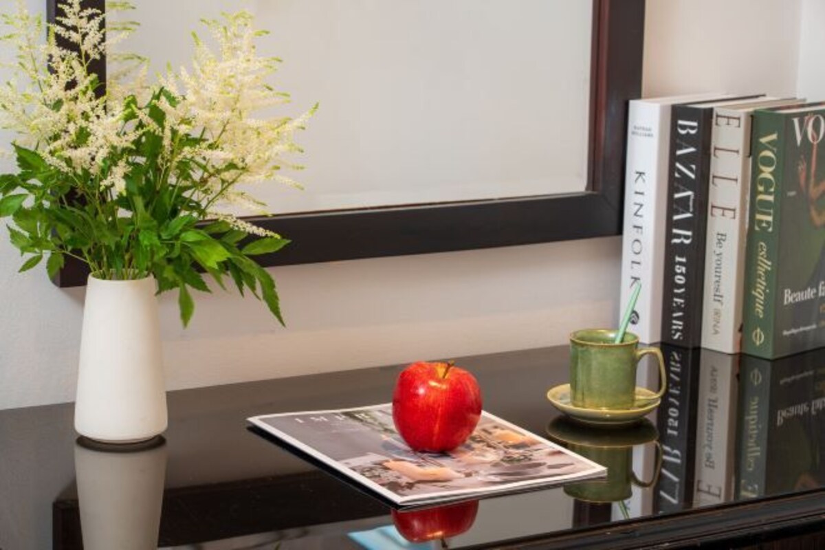 A table with a vase of flowers, a red apple, a green mug, and books.