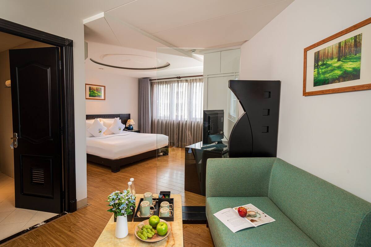Een hotelkamer met een bed, een groene bank, een eettafel met fruit en een vaas, en een schilderij aan de muur.