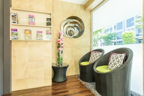 Un vestíbulo moderno con piso de madera, sillas de mimbre con cojines estampados, un estante de pared con revistas y una pieza decorativa de metal.