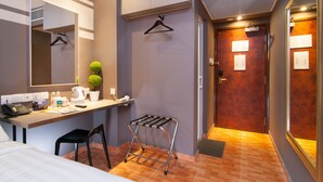 Habitación de hotel con cama, escritorio con teléfono, silla, perchero, planta pequeña en maceta y puerta con espejo.