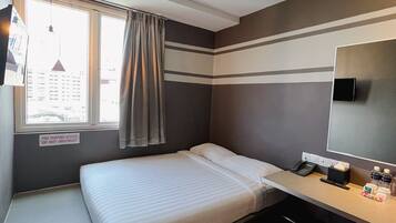 A compact hotel room with a single bed, a desk, and a chair. A window with a view of buildings is visible, and a fire safety sign is mounted on the wall.