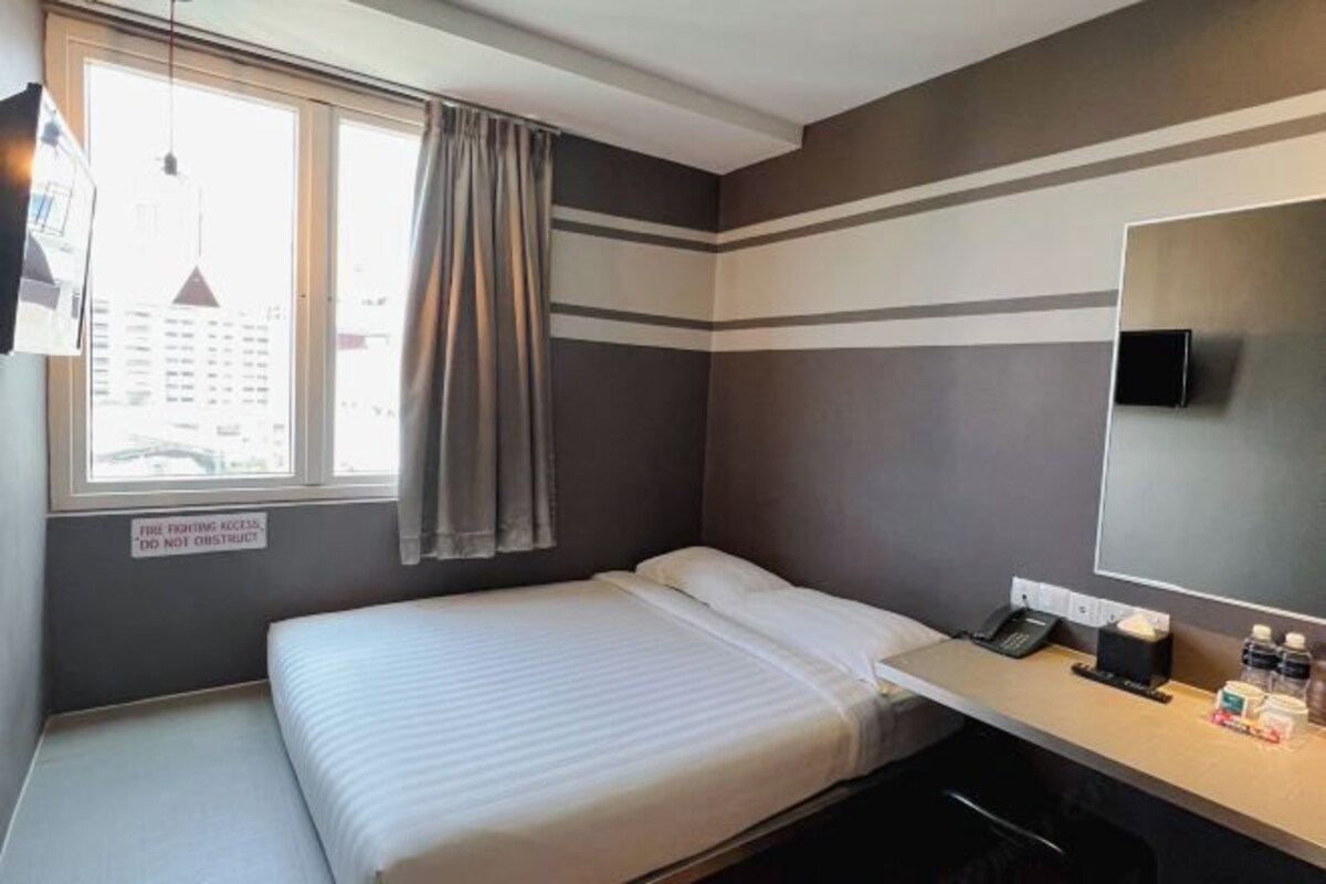 Habitación de hotel con cama, escritorio con teléfono y artículos de aseo, ventana con vistas a edificios, y señalización sobre acceso para bomberos.