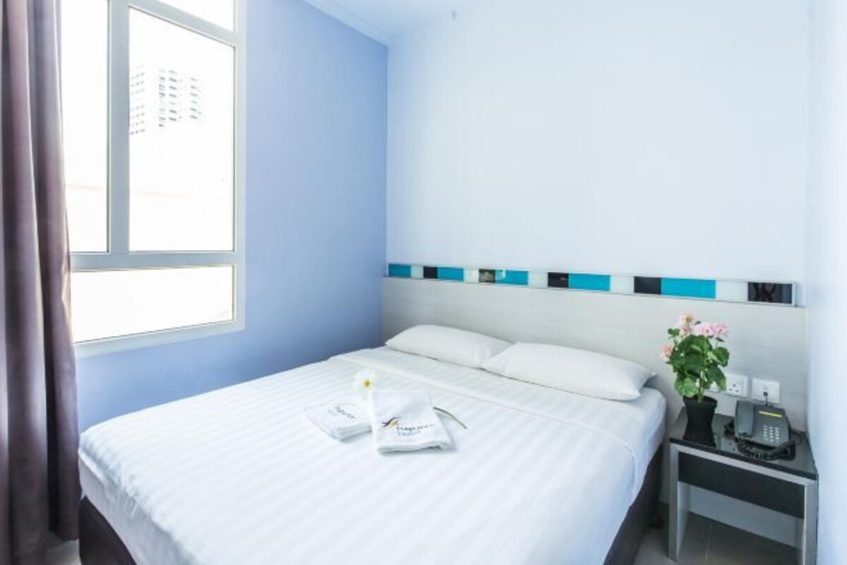 Quarto de hotel com cama, mesa de cabeceira com telefone e janela com cortinas.