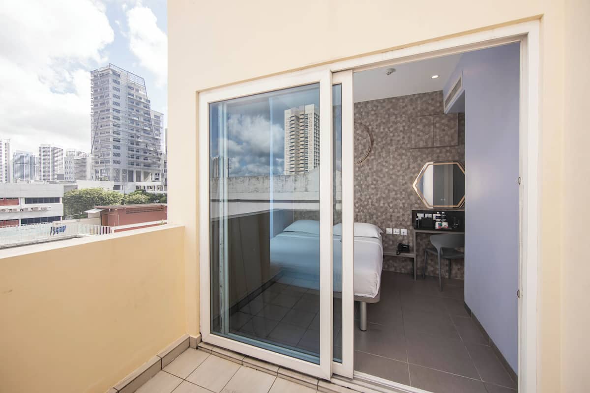 A balcony with a city view, a sliding glass door leading to a bedroom with a bed, a small desk, and a chair.