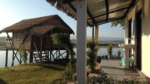 A wooden house with a thatched roof on stilts by a lake, a covered porch with a white column, and a stone pathway leading to a white sink. - Wildlife Adventure Resort (Sauraha)