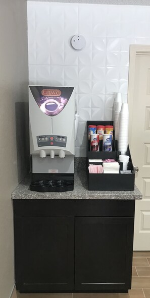 A coffee machine with a control panel and a coffee dispenser, a box of coffee creamer, and a stack of paper cups on a counter. - Red Roof Inn Williamsburg (Williamsburg)