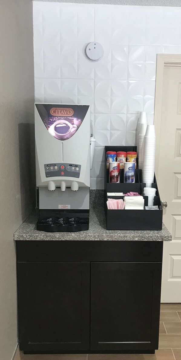A coffee machine with a control panel and a coffee dispenser, a box of coffee creamer, and a stack of paper cups on a counter. - Red Roof Inn Williamsburg (Williamsburg)
