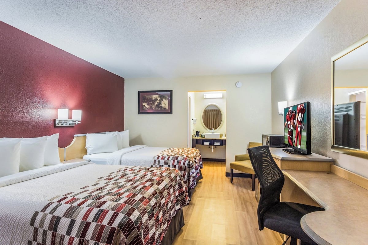 A hotel room with two beds, a desk with a TV, a mirror, and a bathroom with a sink and mirror.
