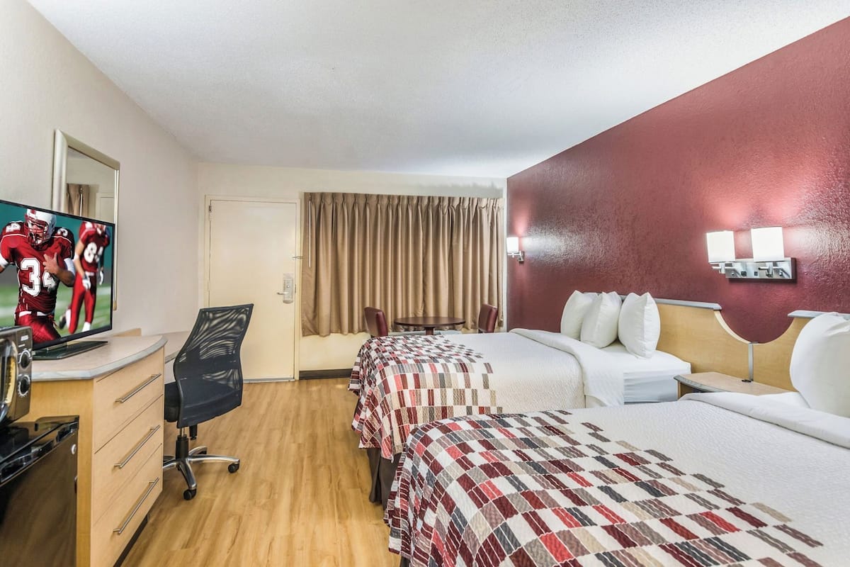 A hotel room with a TV, a desk, two beds, a chair, a window with curtains, and a wall-mounted light fixture.