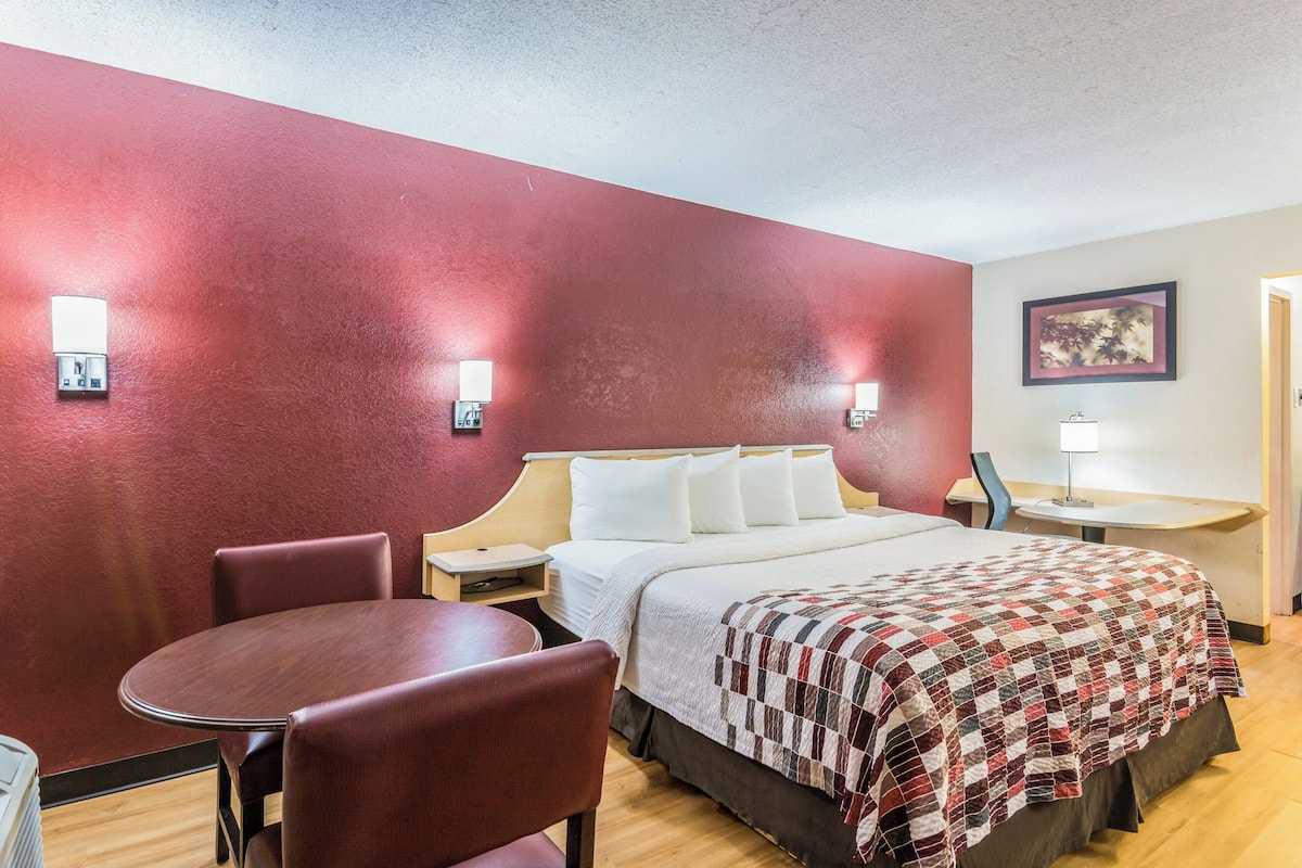 A hotel room with a bed, a table, and two chairs. There is a nightstand with a lamp and a wall-mounted light.