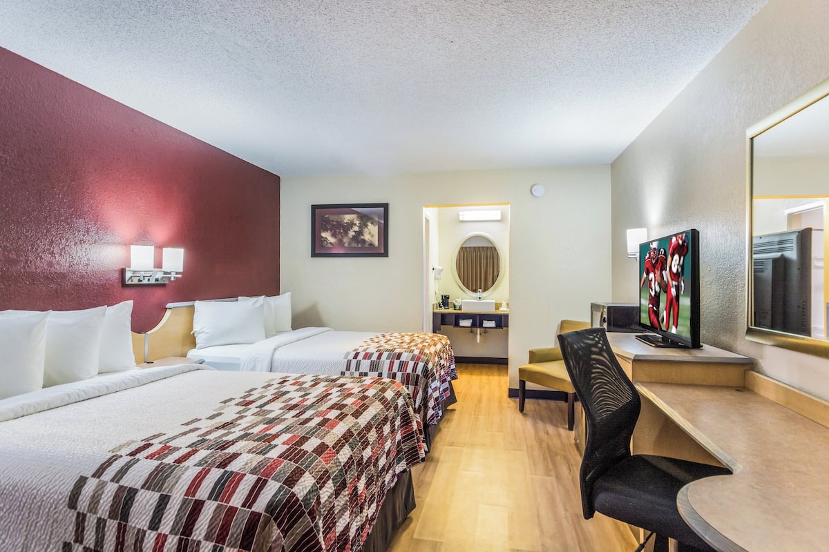 A hotel room with two beds, a desk, a chair, a TV, and a bathroom visible through an open door.